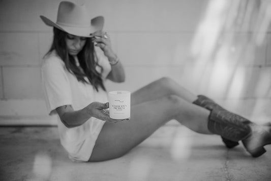A woman in a oversized t shirt, cowgirl boots and cowboy hat tilting her head toward a simple all white candle with black text that says "Somebody's Problem to Love"