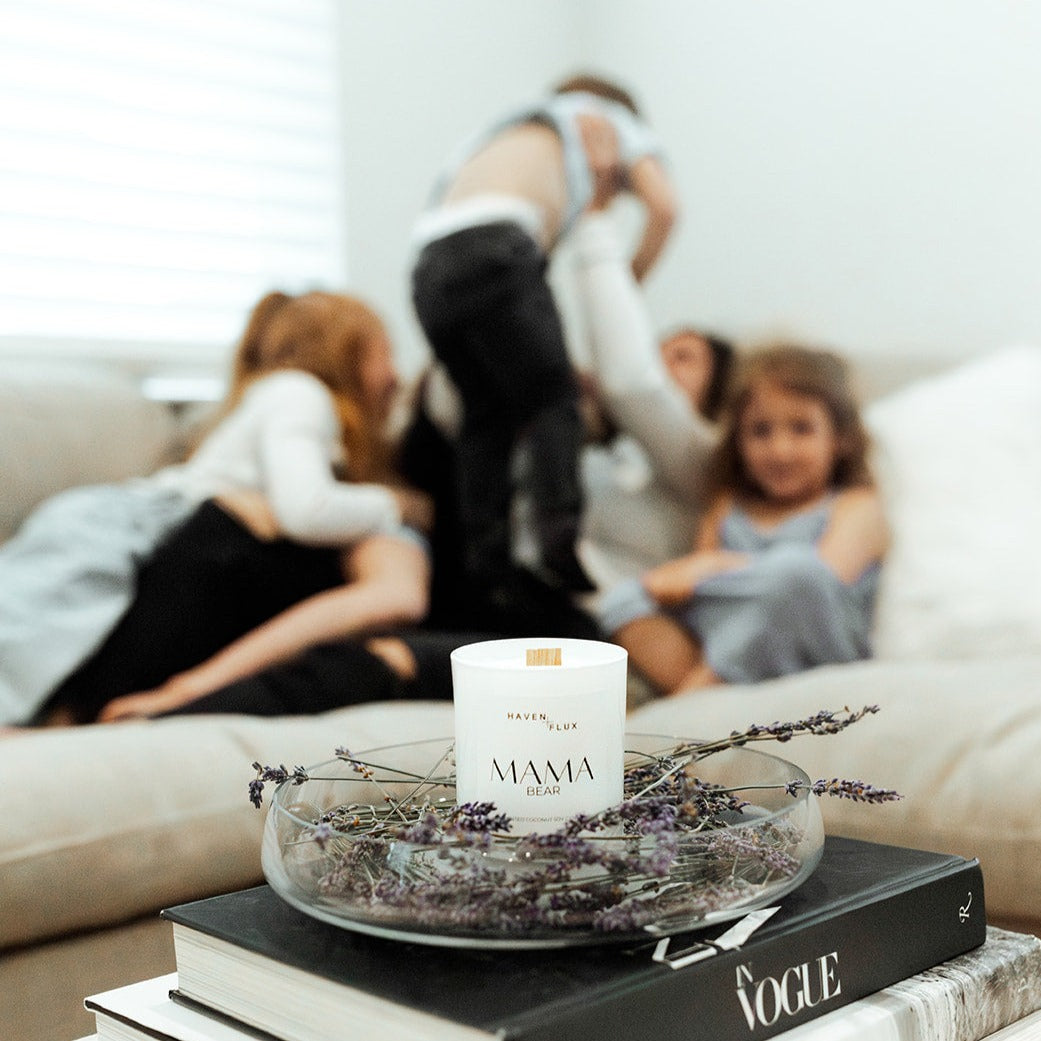 Mama Bear Wooden Wick Candle elegantly placed on a coffee table, with a joyful scene in the background depicting a mother playing with her children. The candle is formulated with luxury, non-toxic ingredients and features scent notes of wild flowers, fresh lavender, and mountain air, creating a serene atmosphere for family moments.